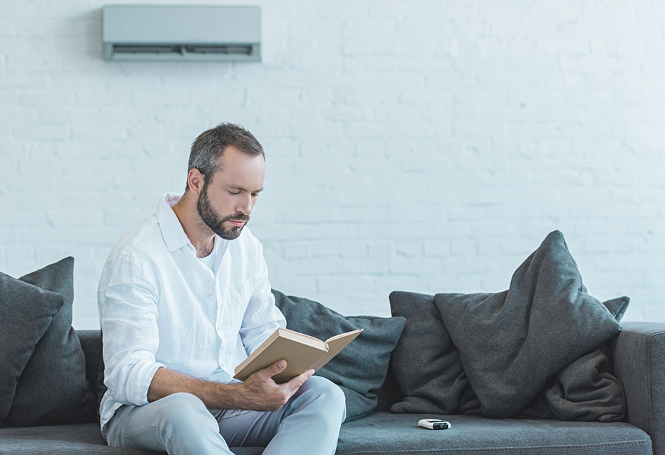 Climaestrat, tus especialistas en las Instalaciones Frigoríficas y de Climatización en Vinaròs / Vinaroz. Hombre leyendo libro en el sofá, con aire acondicionado en la pared.